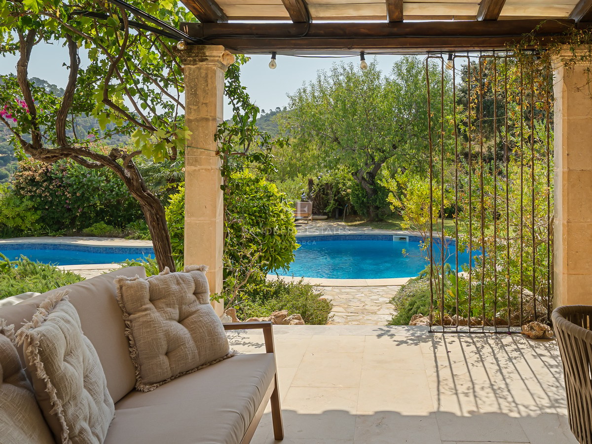 Covered stone terrace with comfortable sofa overlooking a circular blue pool surrounded by lush green gardens.