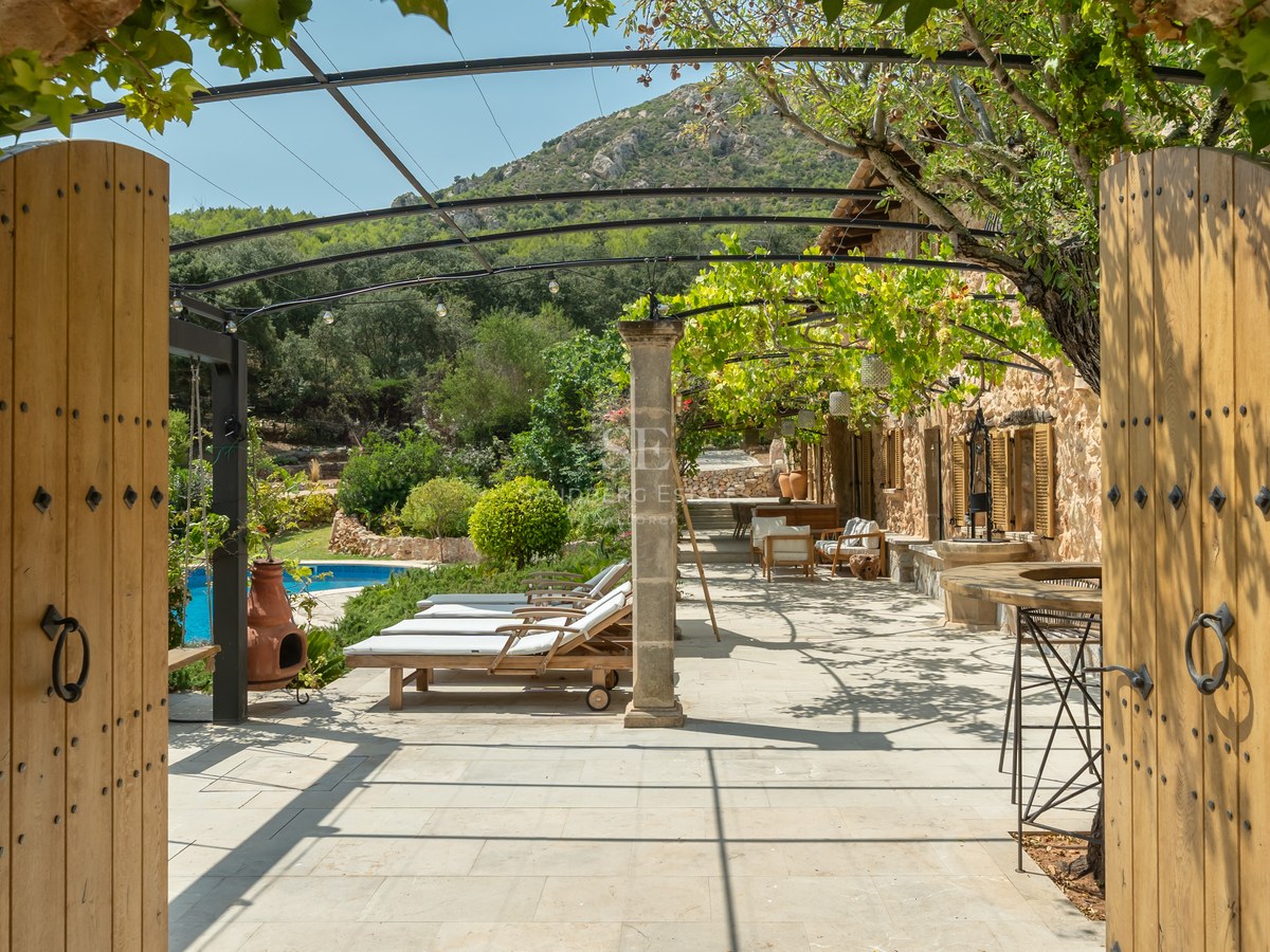 View through open wooden gates of a stone-paved terrace with sun loungers, vine-covered pergola, and pool.