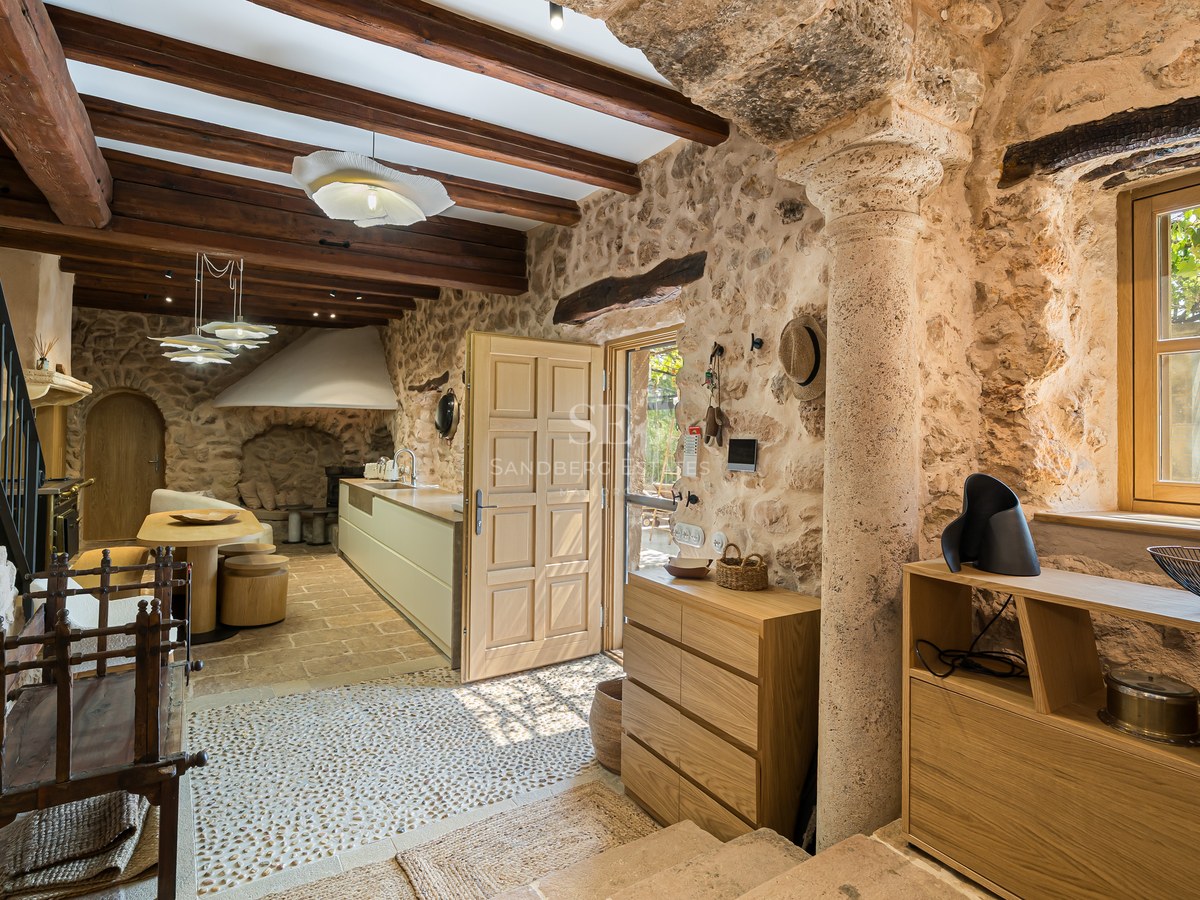 Interior view of a rustic foyer with stone walls, wooden ceiling beams, a stone column, and a view into a modern kitchen.