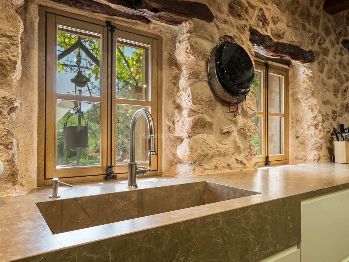 Integrated stone sink under wooden windows in a kitchen featuring exposed stone walls and wooden beams.