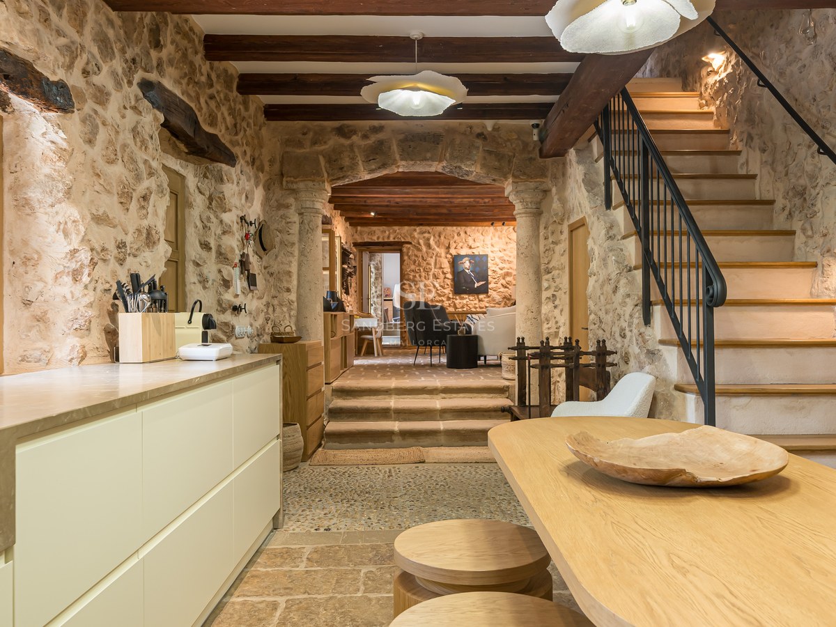 Rustic kitchen with exposed stone walls, wooden beams, a modern island, and a stone staircase leading upstairs.