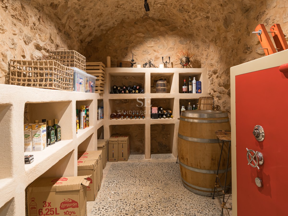 Bodega rústica con techos de piedra abovedados, estanterías de obra, mesa de barril y una caja fuerte roja.