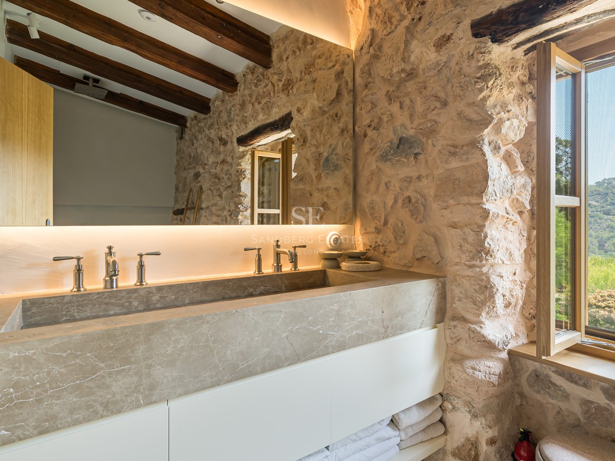 Modern integrated natural stone double vanity against a rustic stone wall with timber beams and a scenic window view.