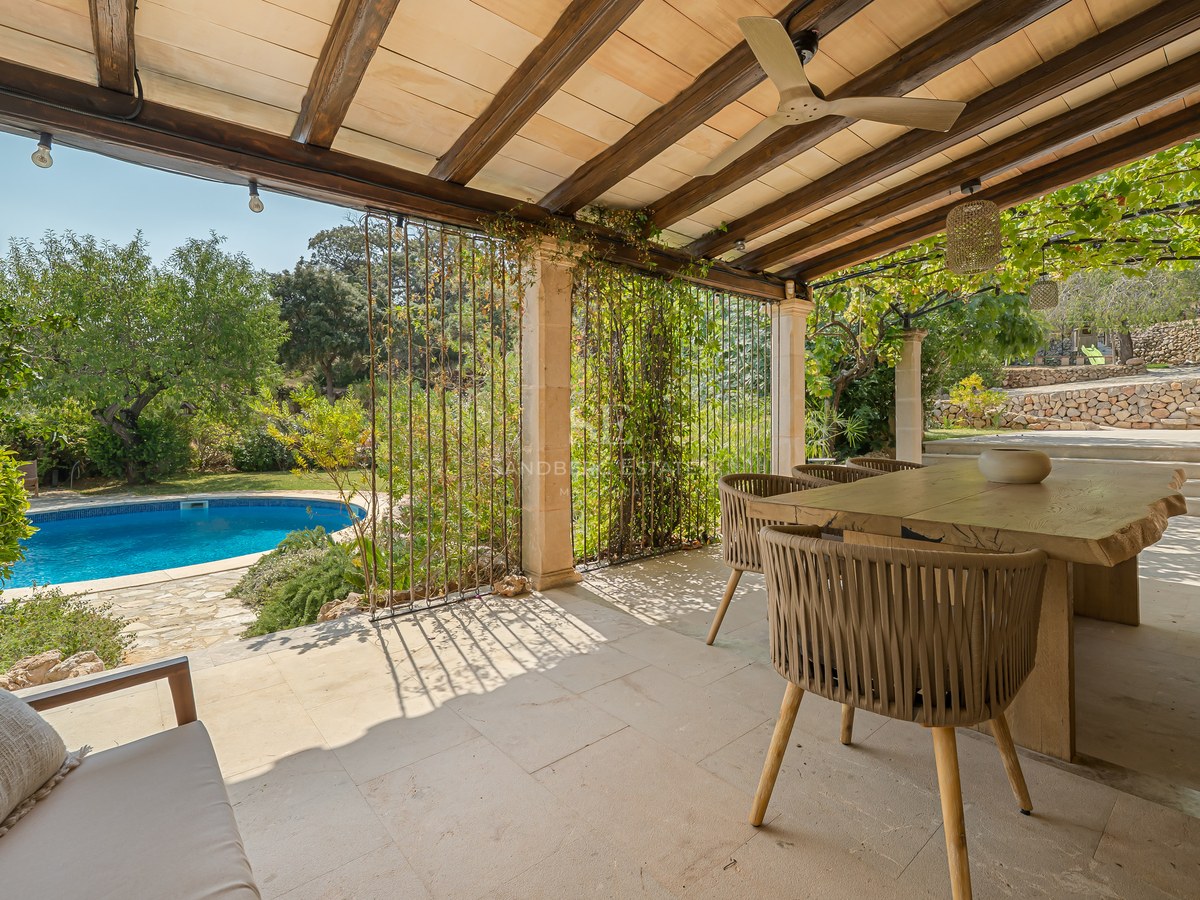 A covered stone terrace with wooden beams, a rustic dining table, and views over a lush garden and turquoise pool.