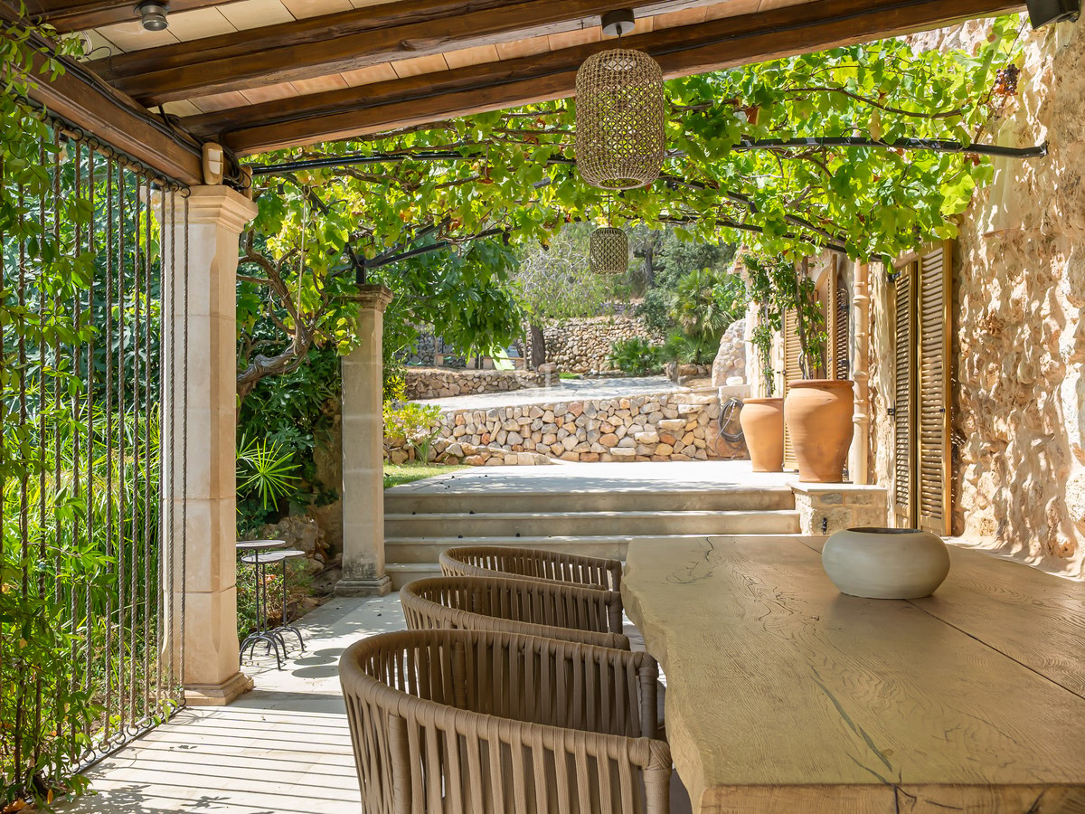 Outdoor dining area under a vine-covered pergola with a live-edge wooden table and natural stone walls.