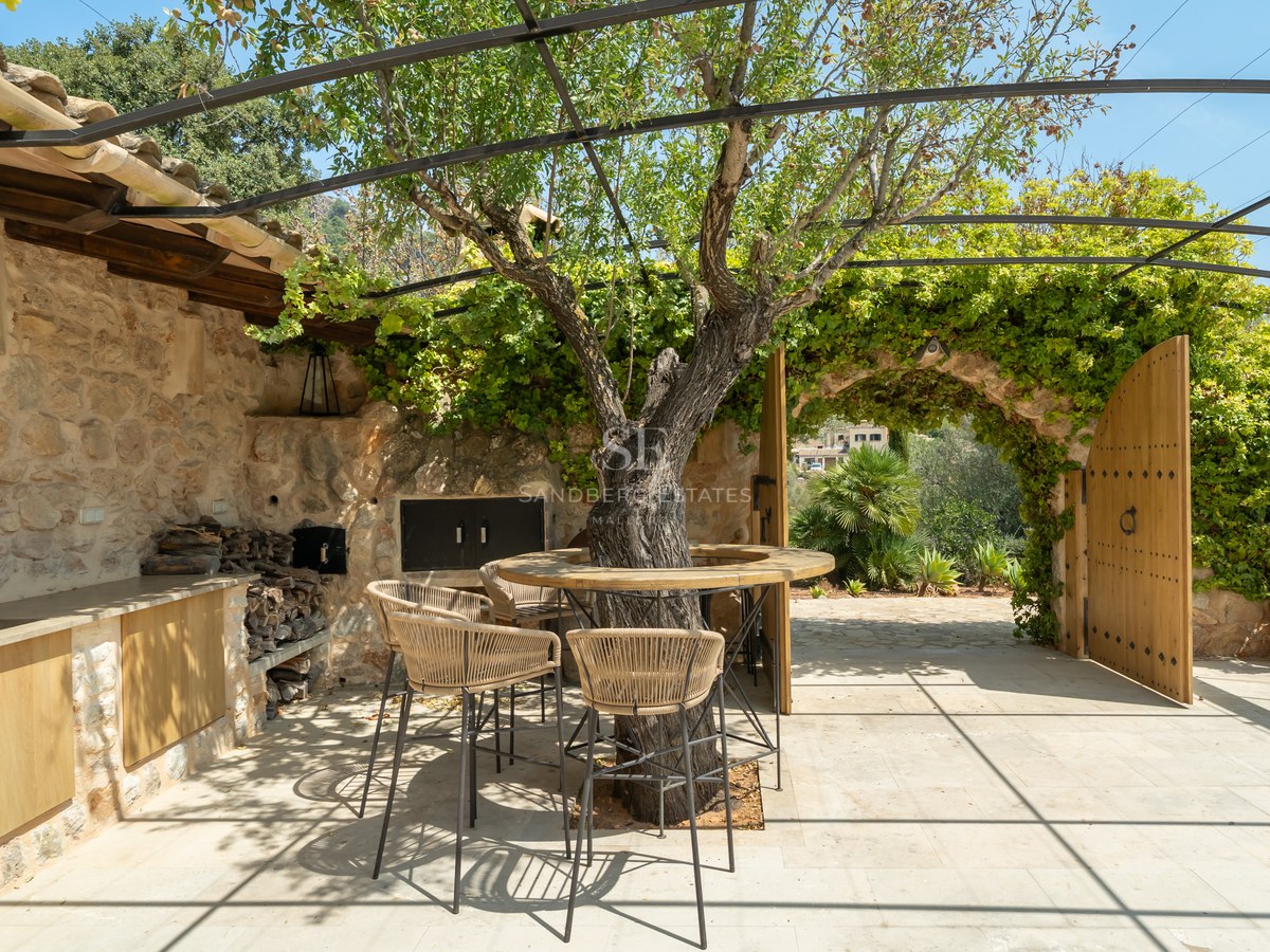 Rustic stone terrace with outdoor kitchen, integrated tree bar table, and arched wooden gates under a metal pergola.