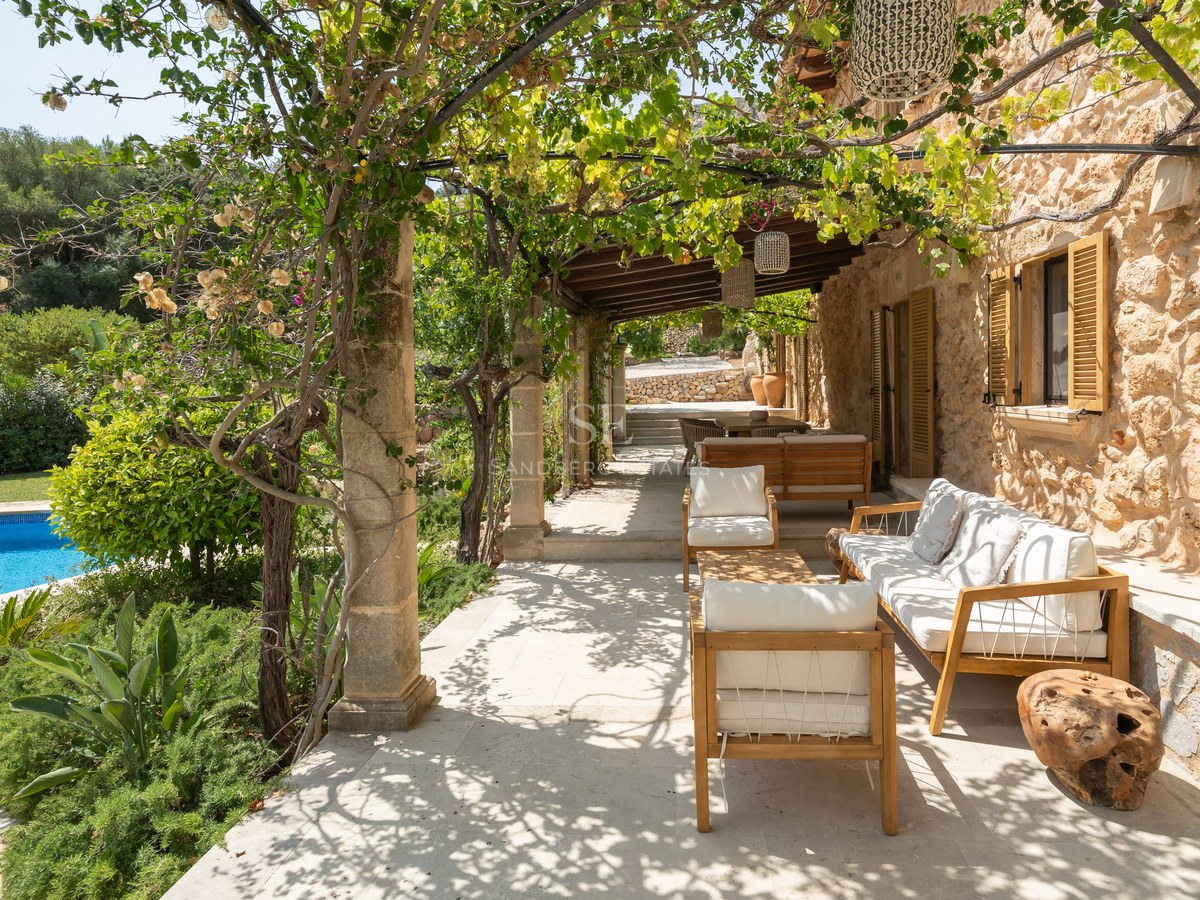 Sun-dappled stone terrace with white cushioned wooden seating, lush vines, and a blue pool in the background.