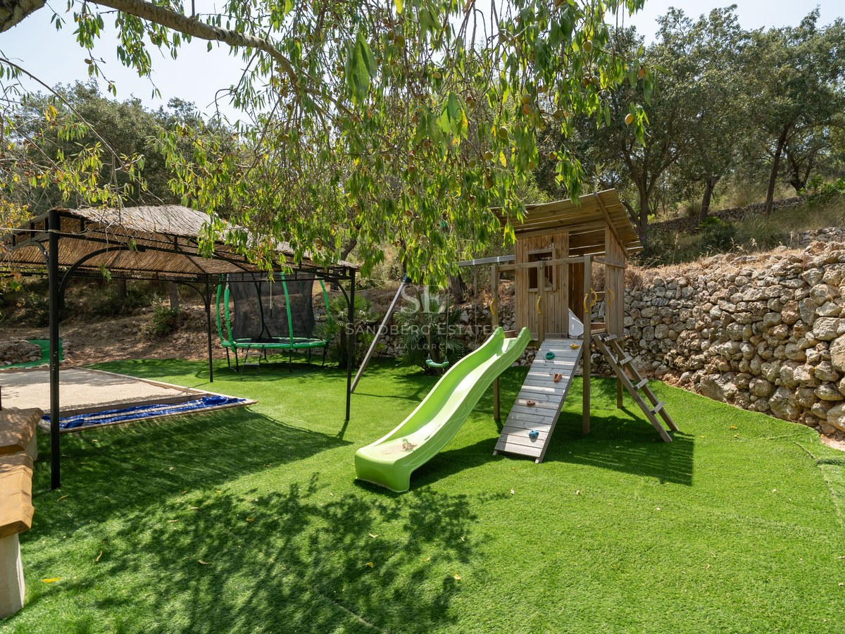 Children's wooden playhouse with a green slide, trampoline, and thatched gazebo on artificial grass bordered by a stone wall.