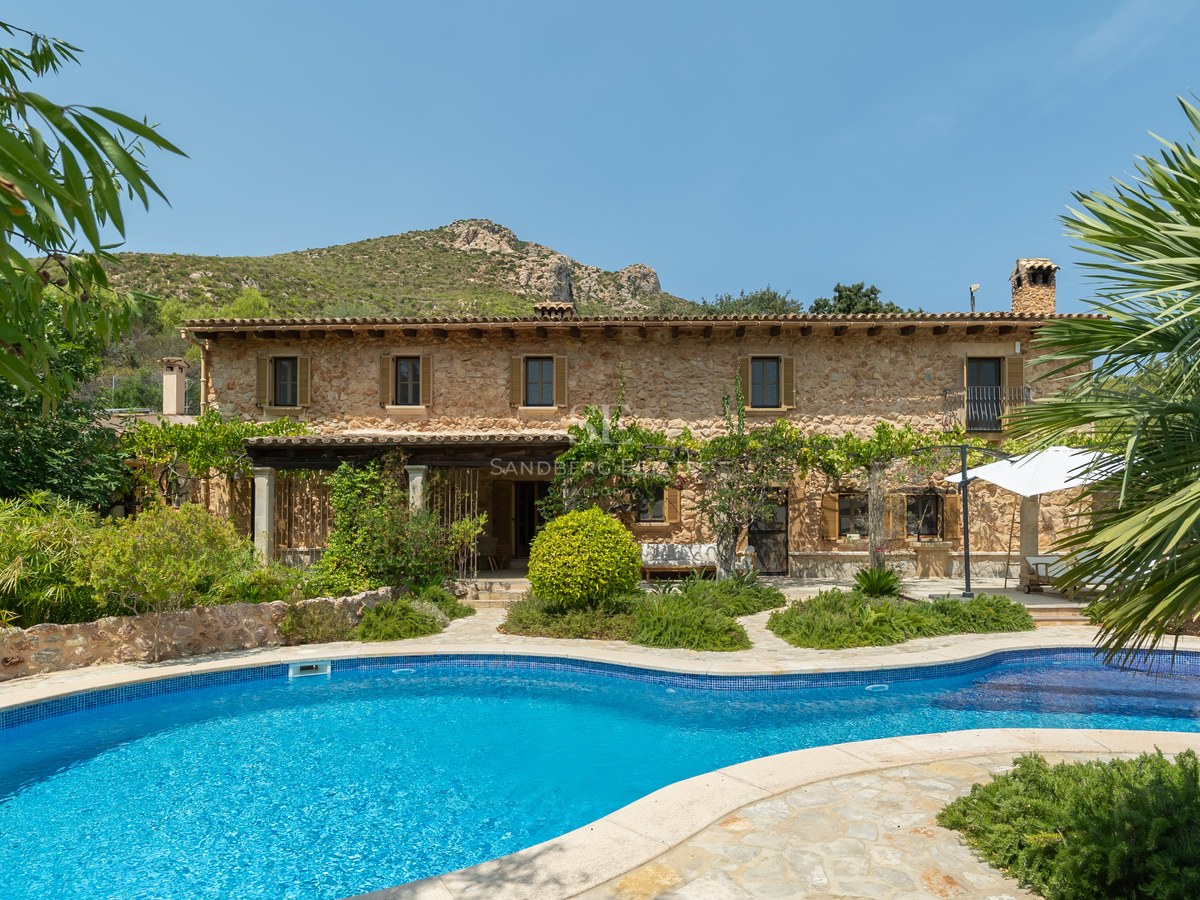 Large natural stone villa with a curved blue swimming pool, lush gardens, and mountain views under a clear sky.