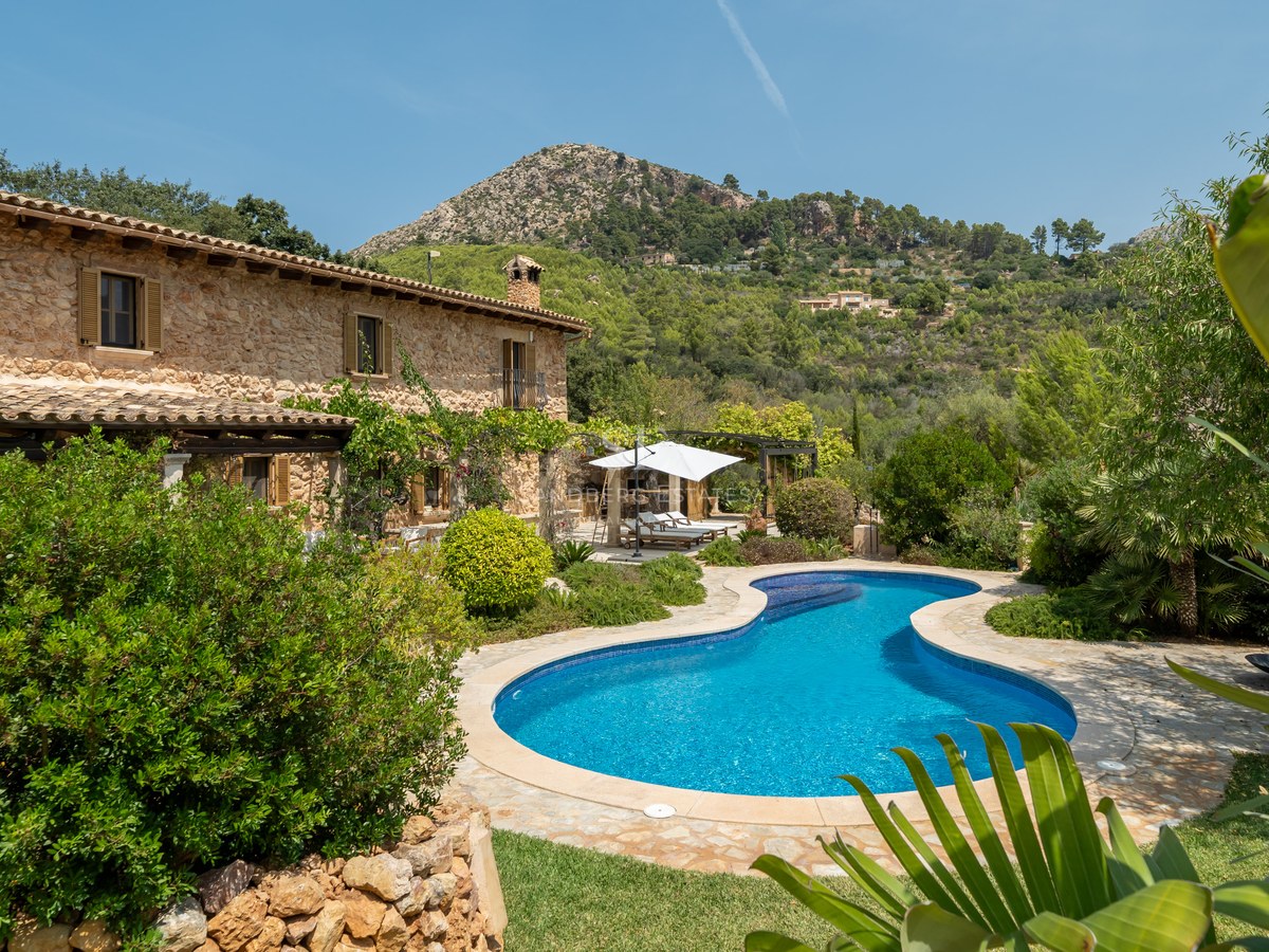 Luxury stone farmhouse with a curved blue swimming pool, lush green gardens, and a scenic mountain backdrop.