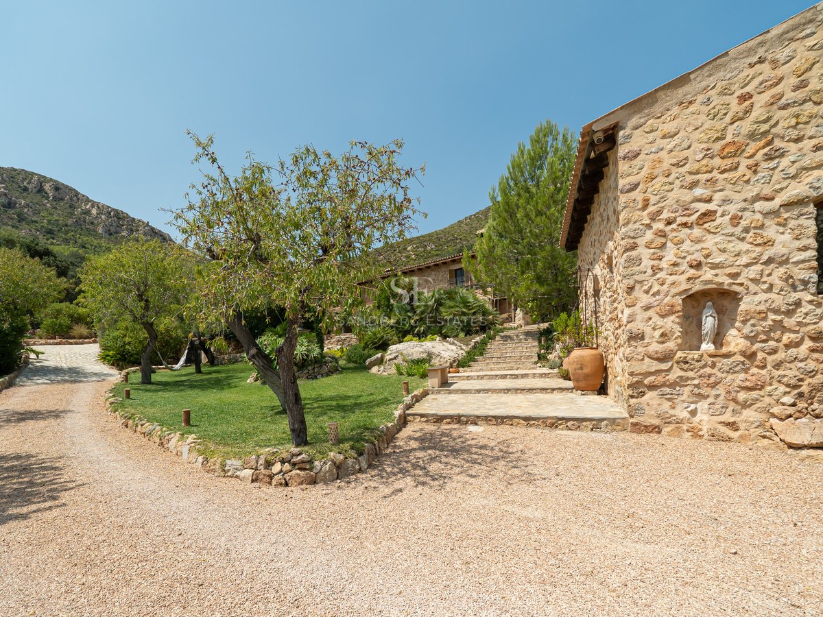 Natural stone villa exterior with gravel driveway, manicured gardens, and mountain backdrop under a clear blue sky.