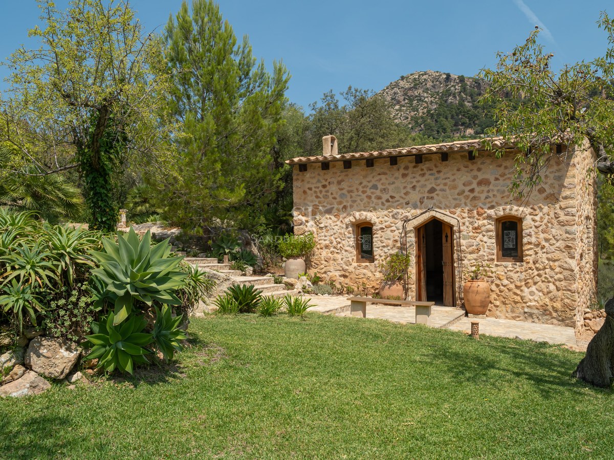 Stone cottage with terracotta roof nestled in a Mediterranean garden against a mountain backdrop.