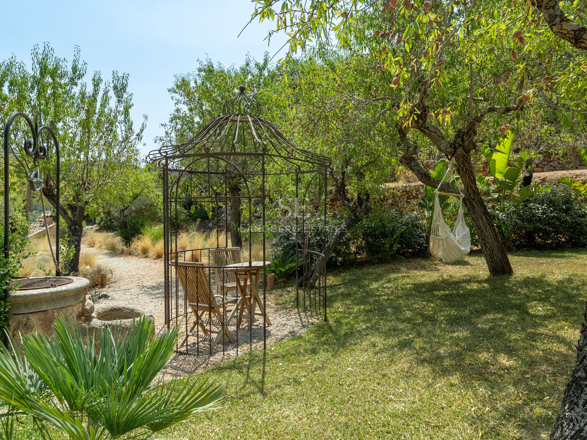 Wrought iron gazebo and stone well in a lush Mediterranean garden with a white hammock hanging from a tree.