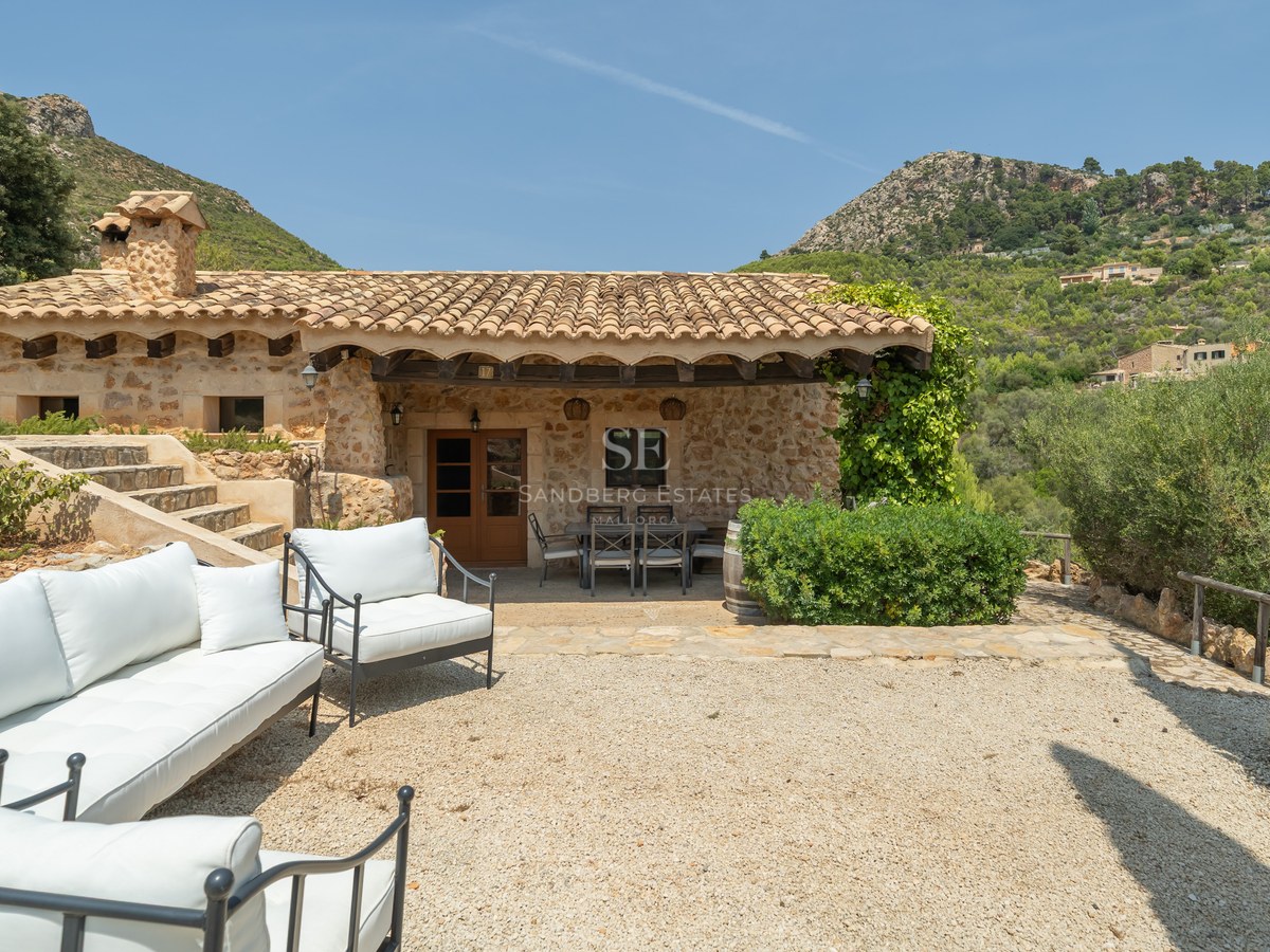 A rustic stone villa with a gravel terrace featuring white sofas and a covered dining area against a mountain backdrop.