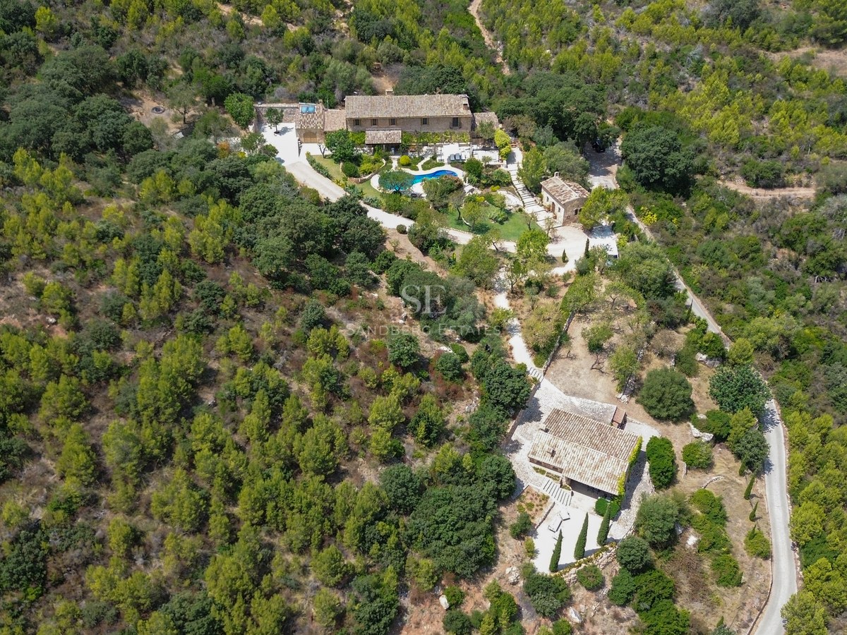 Vista aérea de una gran finca de piedra con piscina y casa de invitados rodeada de un denso bosque mediterráneo.