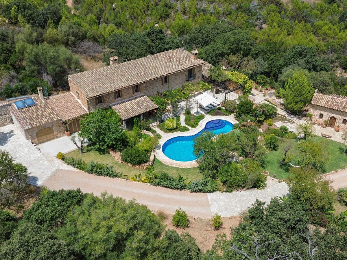 Aerial shot of a stone villa with a blue pool, guest house, and gardens surrounded by green forest.