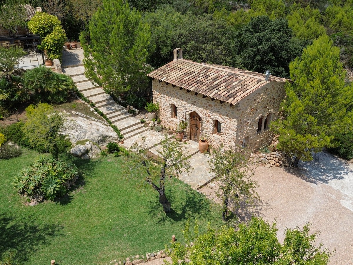 Vista aérea de una casita de piedra rústica con tejado de terracota rodeada de jardines y árboles mediterráneos.