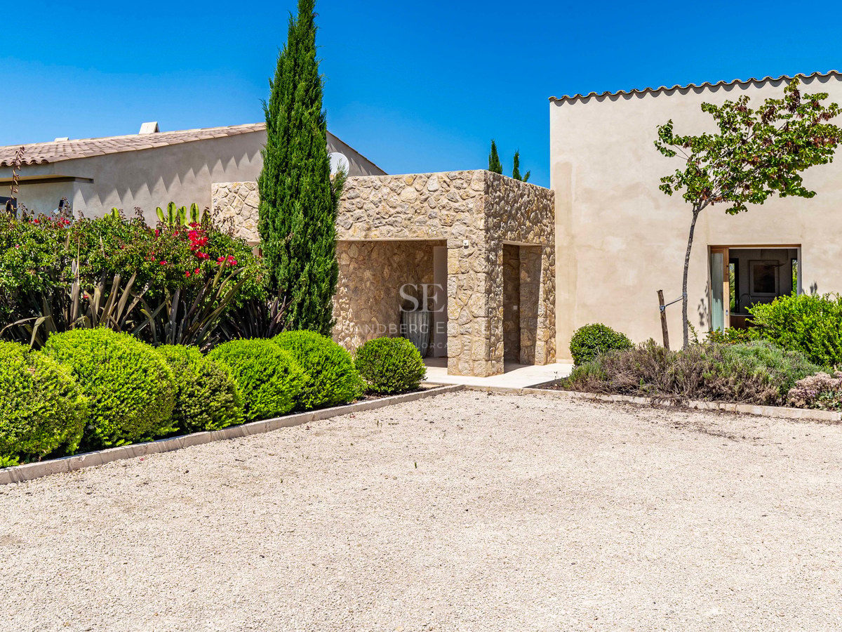 Entrée de villa en pierre avec allée de gravier et arbustes verts sous un ciel bleu.