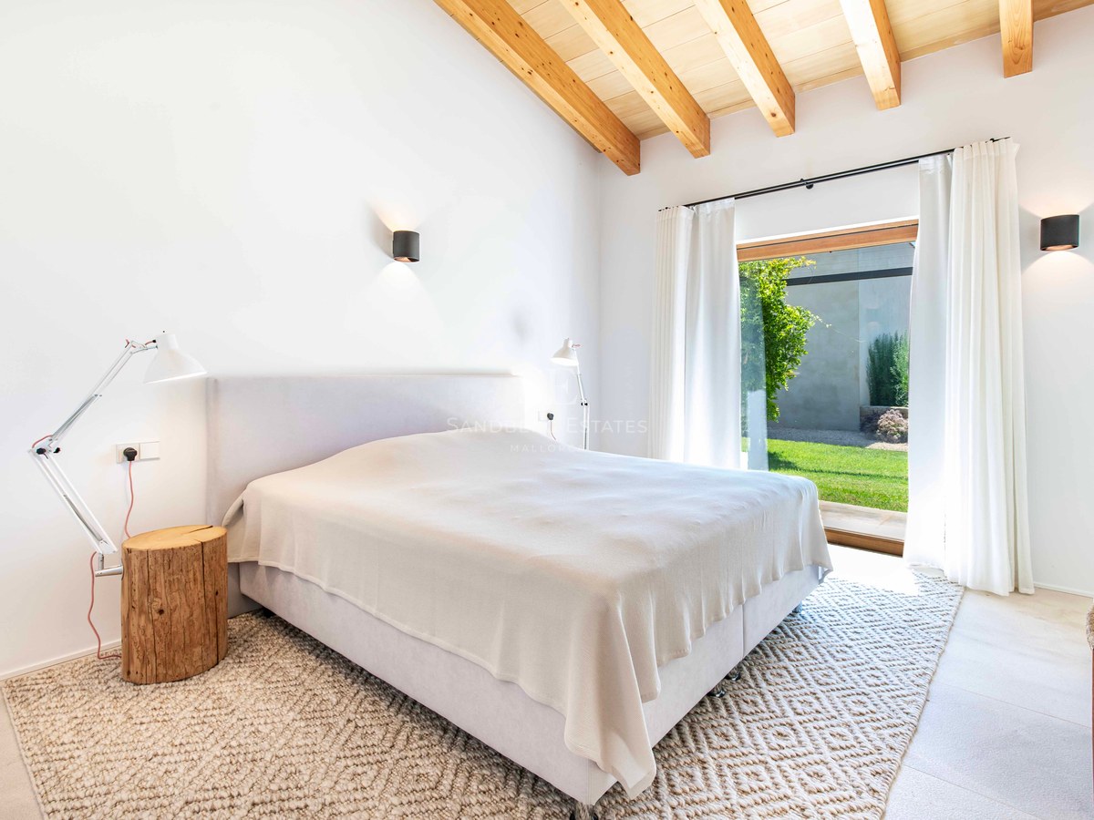 Chambre lumineuse avec plafond voûté en bois, murs blancs, grande baie vitrée donnant sur un jardin et tapis texturé.