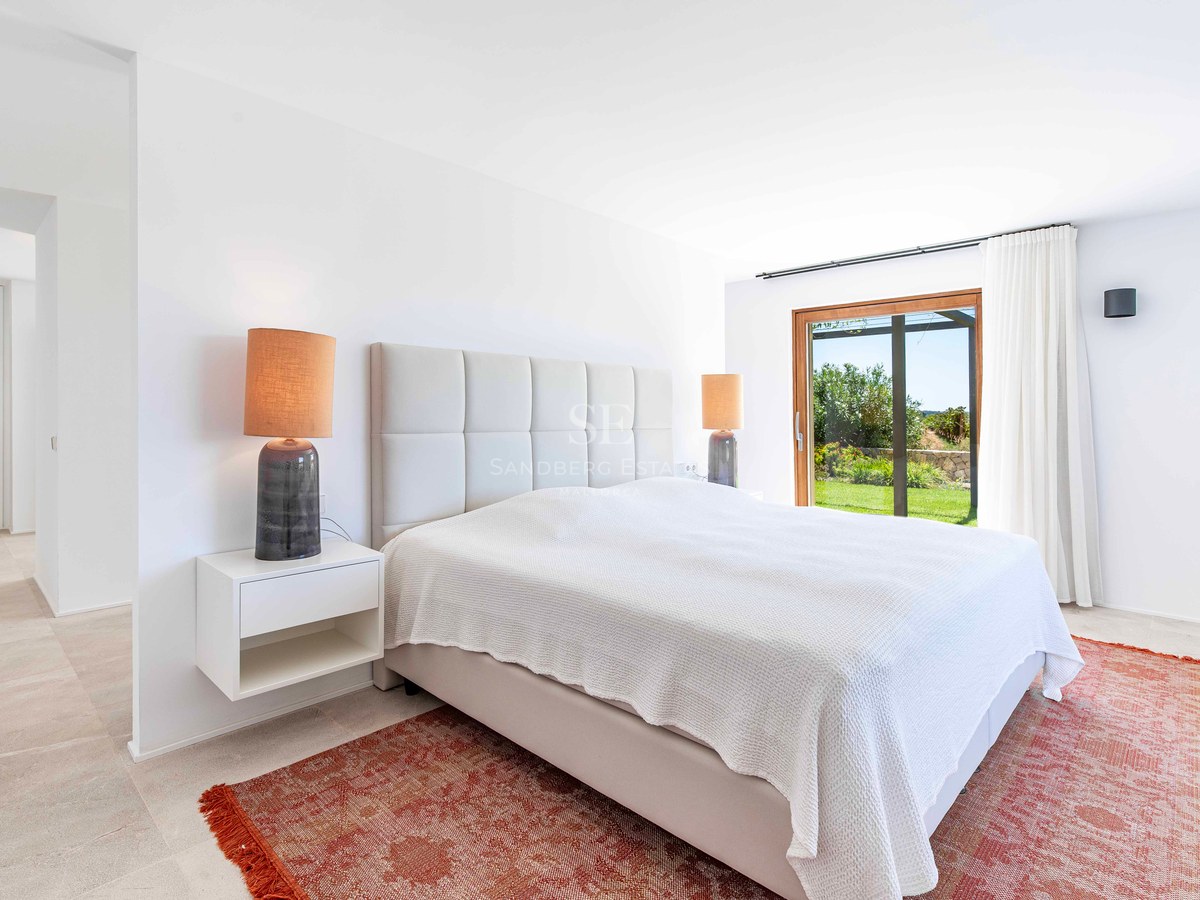 Chambre moderne lumineuse avec grand lit, tapis terre cuite et baies vitrées ouvrant sur un jardin verdoyant.
