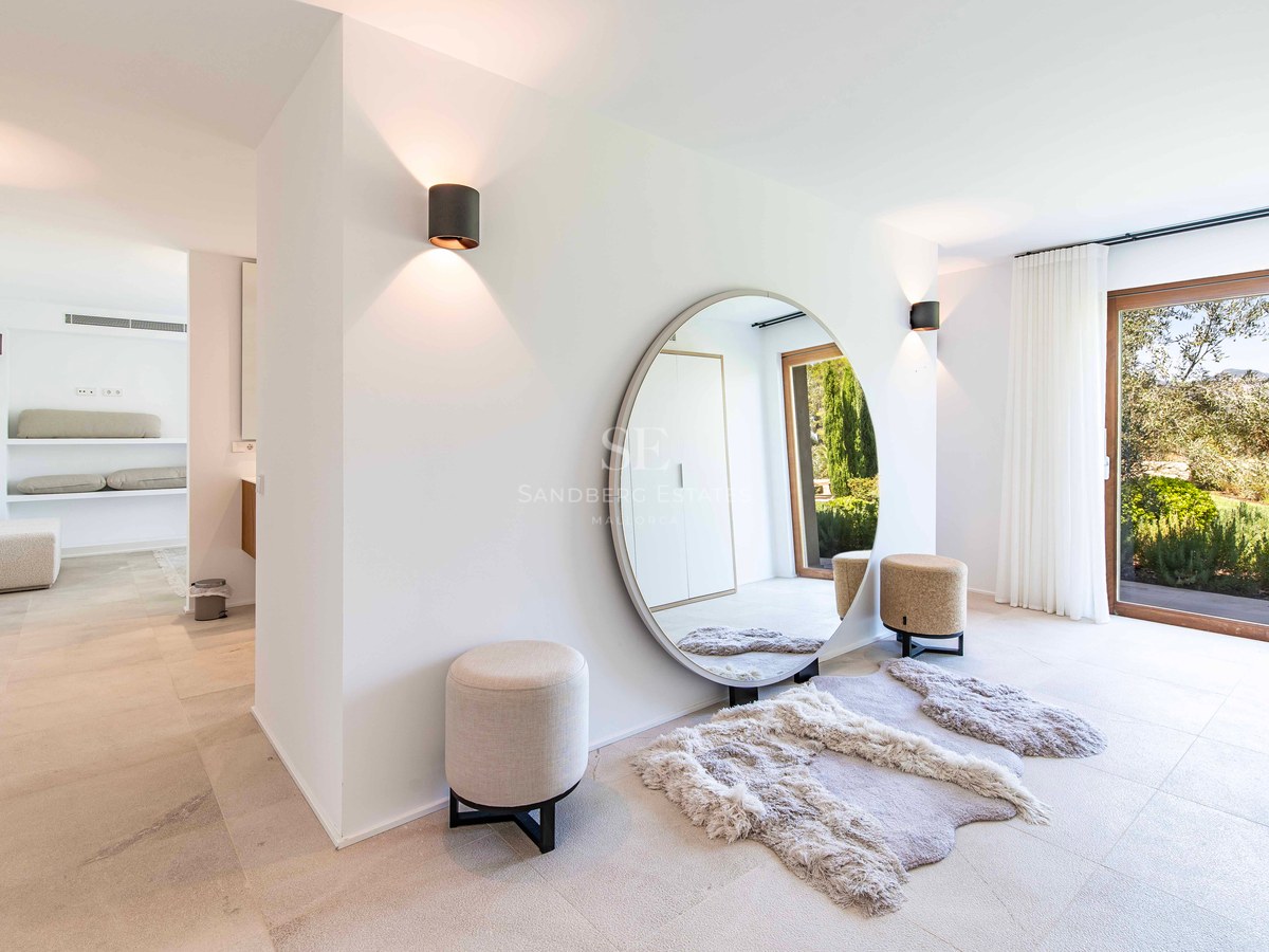 Couloir minimaliste avec grand miroir circulaire, poufs crème et vue sur un jardin via une porte vitrée.