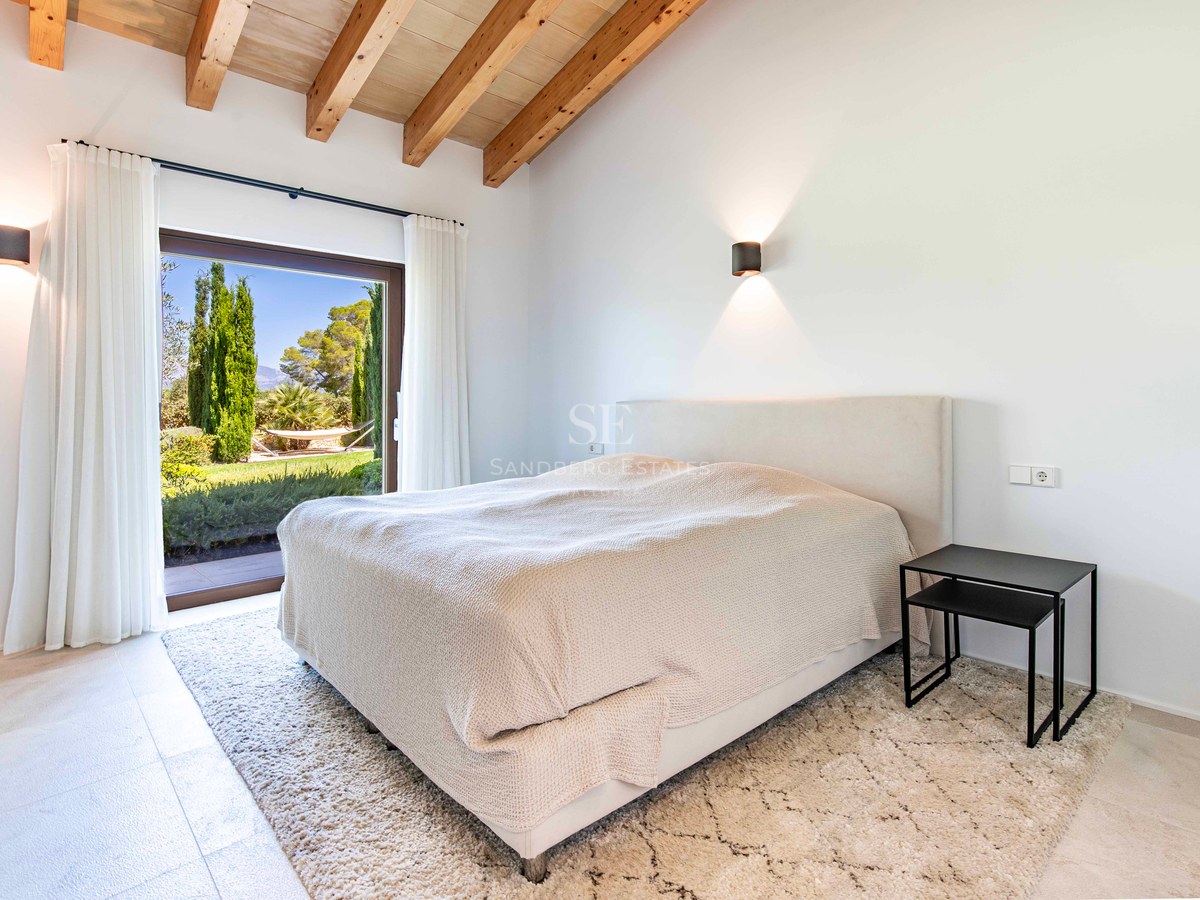 Chambre moderne avec poutres apparentes, lit blanc et grande baie vitrée ouvrant sur un jardin luxuriant.