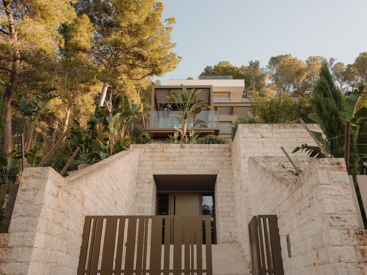 Vista en contrapicado de una villa moderna con muros de piedra natural, balcones de cristal y pinos al atardecer.