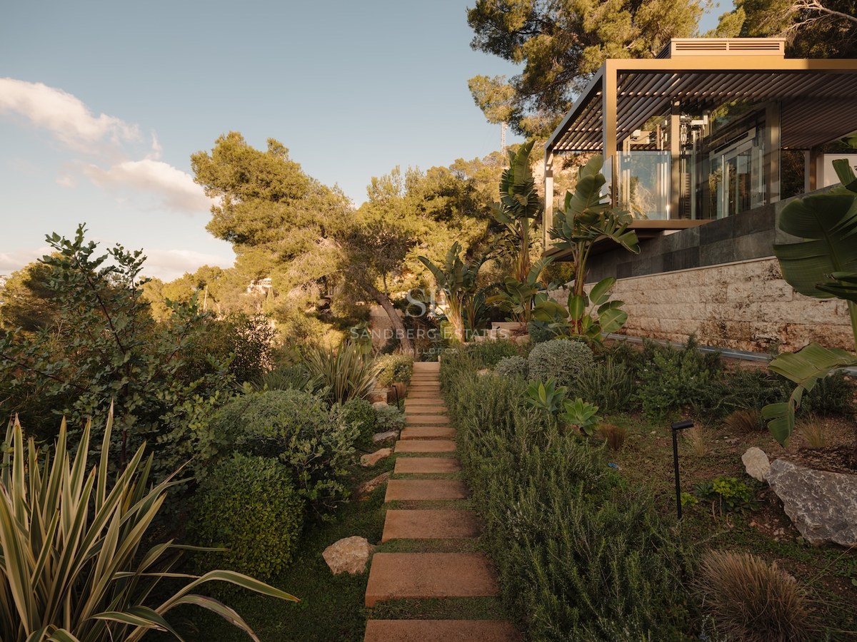 Sendero de piedra entre vegetación mediterránea hacia un pabellón de cristal moderno con pérgola metálica.