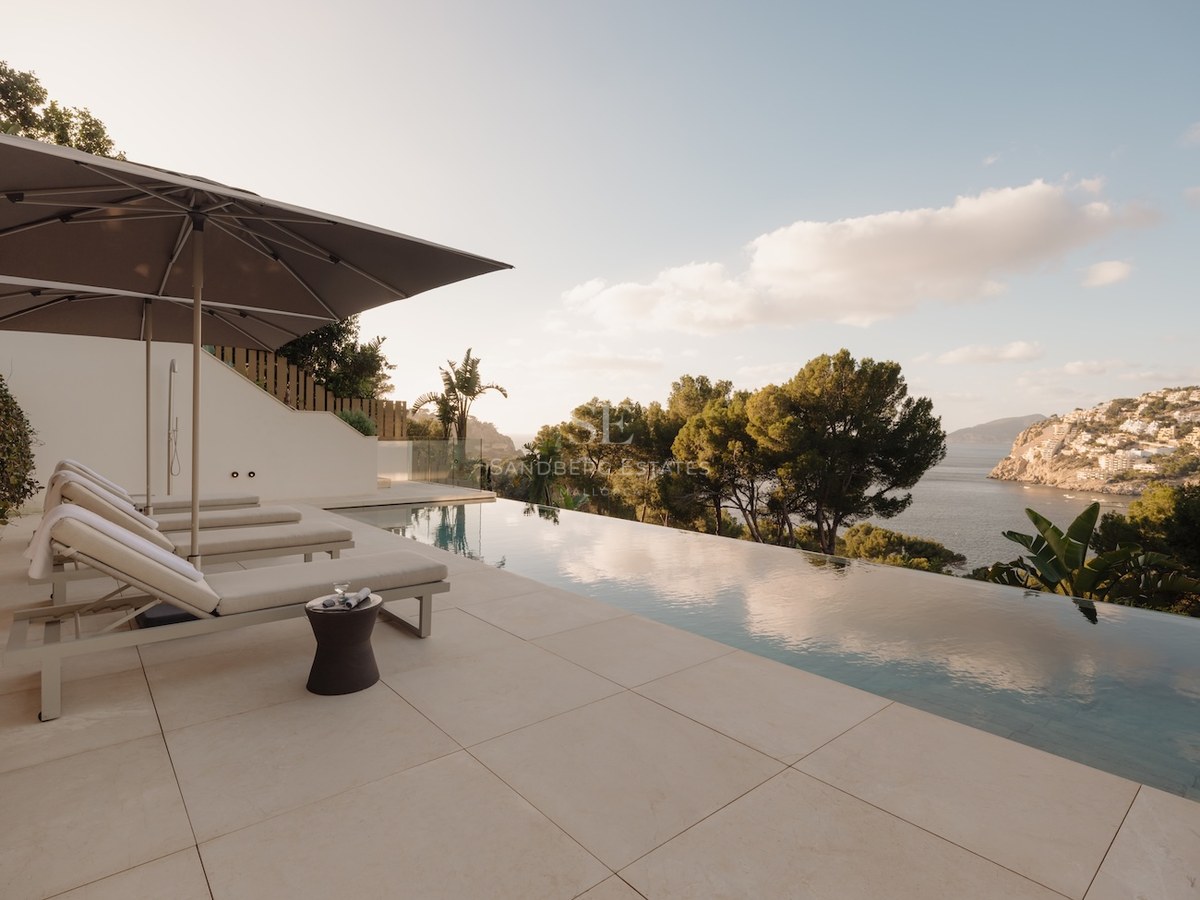 Piscina infinita de lujo y terraza de piedra con hamacas frente a una bahía mediterránea con pinos y agua azul.