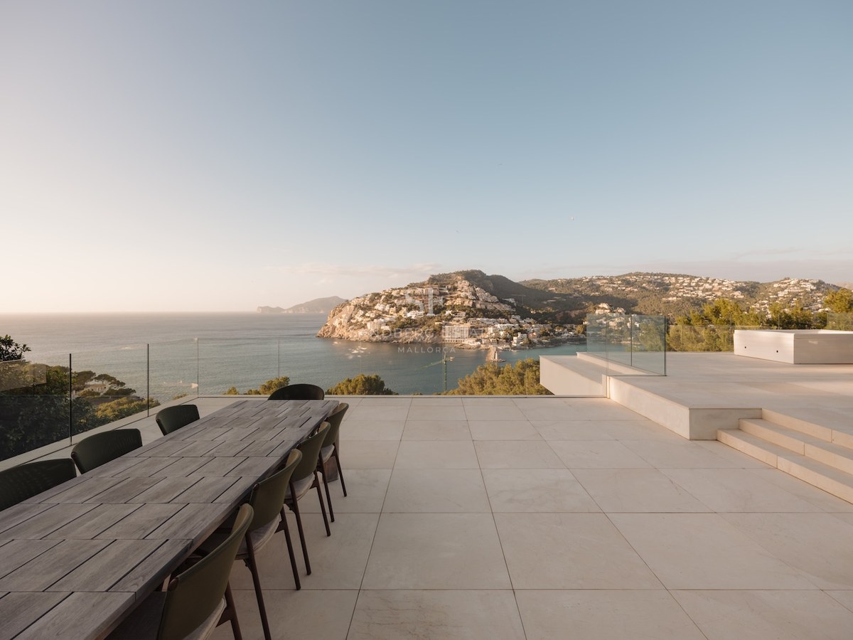 Terraza de piedra elevada con mesa de comedor de madera con vistas a una bahía mediterránea y montañas al atardecer.