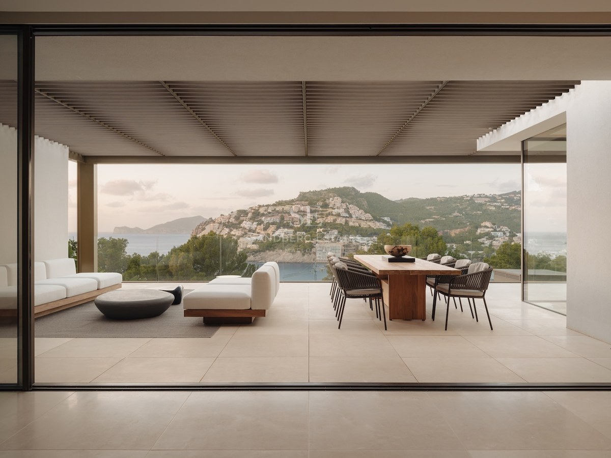 Terraza moderna cubierta con mesa de comedor de madera, sofás blancos y vistas al mar tras barandillas de cristal.