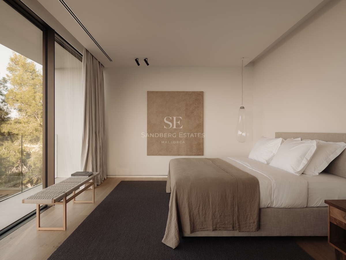 Dormitorio minimalista con ventanales de suelo a techo, suelo de madera, ropa de cama de lino y vistas a los árboles.