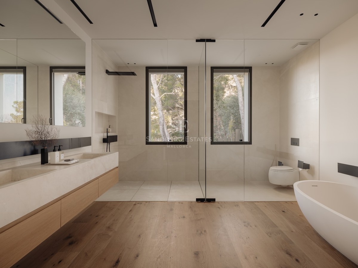 Baño moderno con suelos de roble, lavabo doble de piedra, bañera exenta y ducha frente a ventanales con pinos.