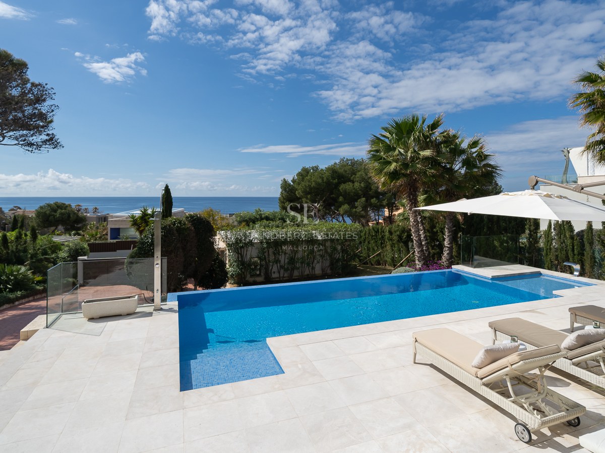 Blue outdoor swimming pool with sun loungers and light stone terrace overlooking the sea and palm trees.