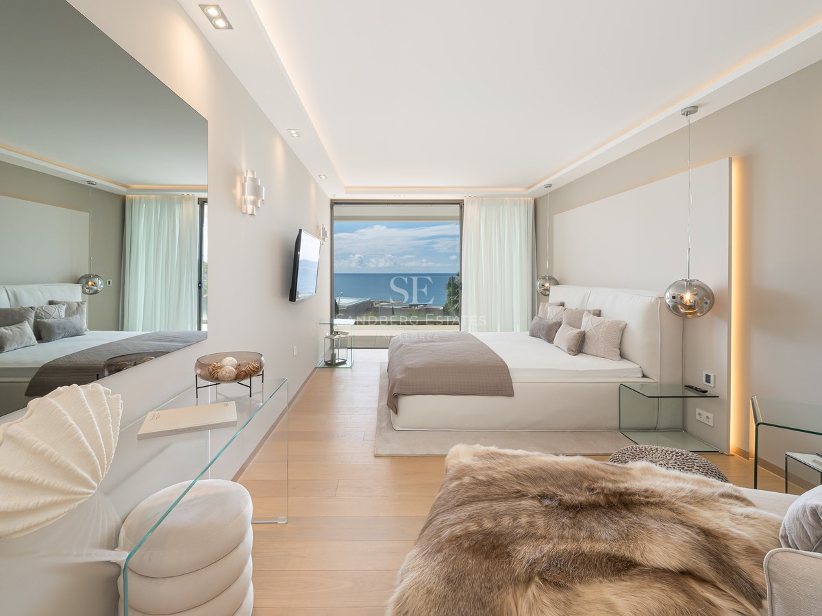Luxury bedroom with white bed, floor-to-ceiling glass doors overlooking the ocean, glass furniture, and light oak floors.