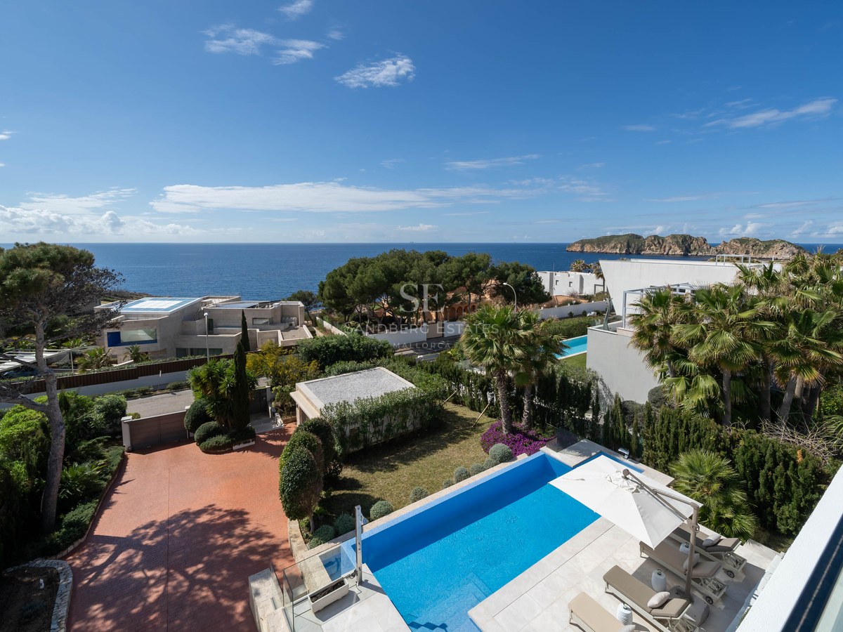 Elevated view of a blue swimming pool, sun deck with loungers, Mediterranean gardens, and sea views under a clear sky.