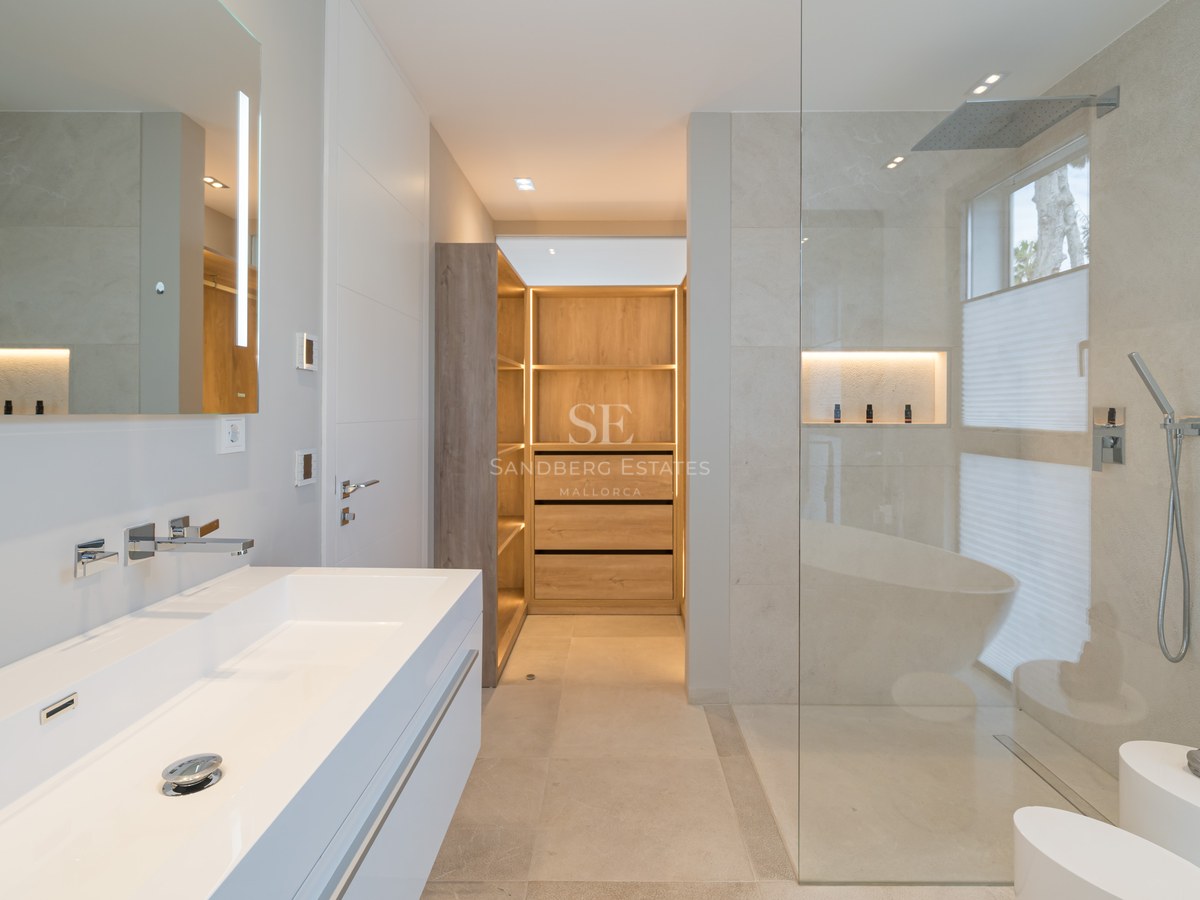 High-end modern bathroom featuring a white double vanity, glass rain shower, and an illuminated wooden walk-in closet.