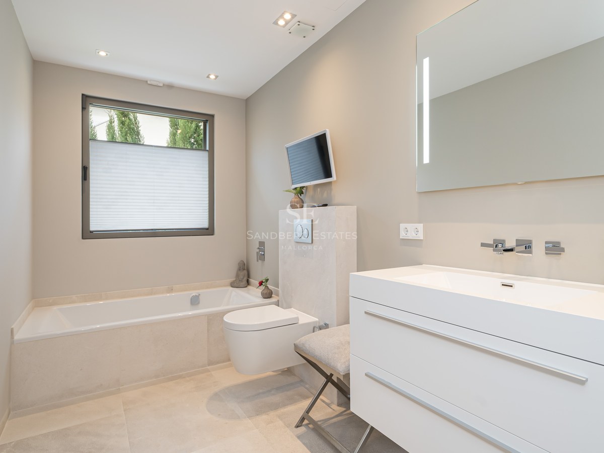 Minimalist bathroom featuring a white vanity, a built-in bathtub, a wall-hung toilet, and a wall-mounted television.
