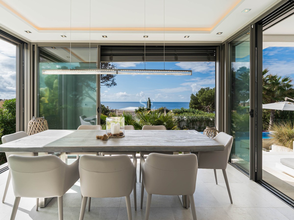 Modern dining room featuring a large wooden table and grey chairs, surrounded by glass walls overlooking the sea.