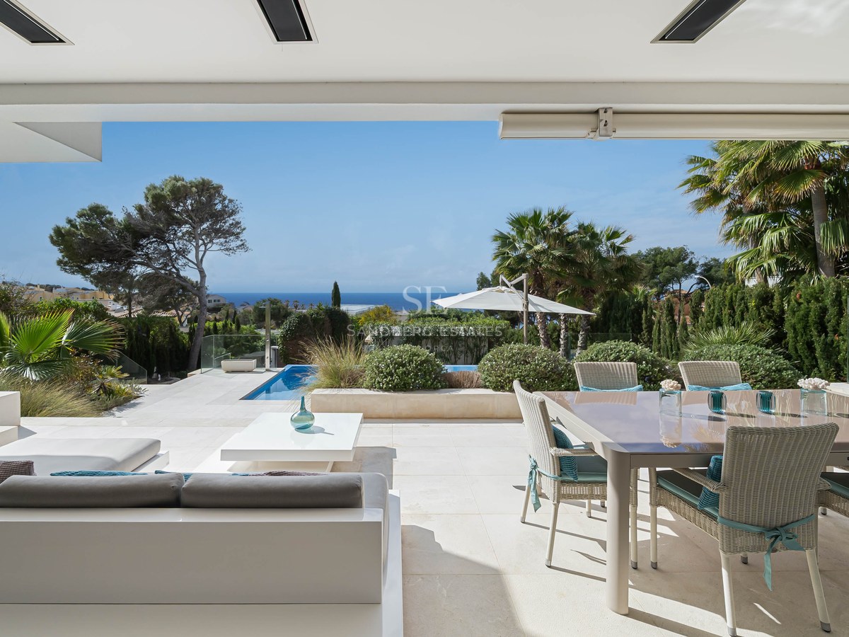 Spacious luxury terrace with white lounge seating, wicker dining set, stone floors, and a view of the pool and sea.