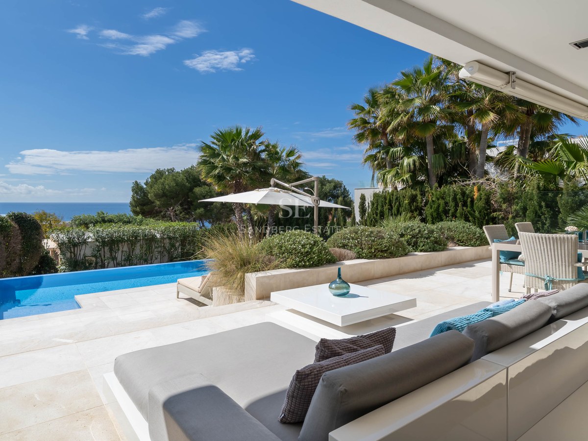 Luxury terrace with white lounge furniture, stone flooring, infinity pool, and palm trees overlooking the sea.