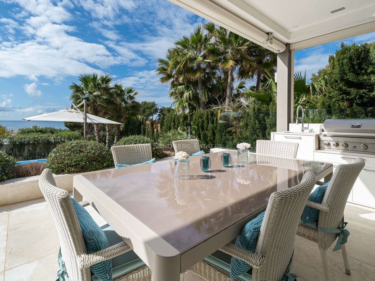 Outdoor dining table and integrated BBQ kitchen on a stone terrace overlooking the sea and palm trees.