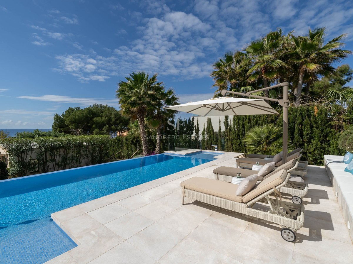 A sun-drenched terrace featuring an infinity pool, beige sun loungers under an umbrella, and distant sea views.