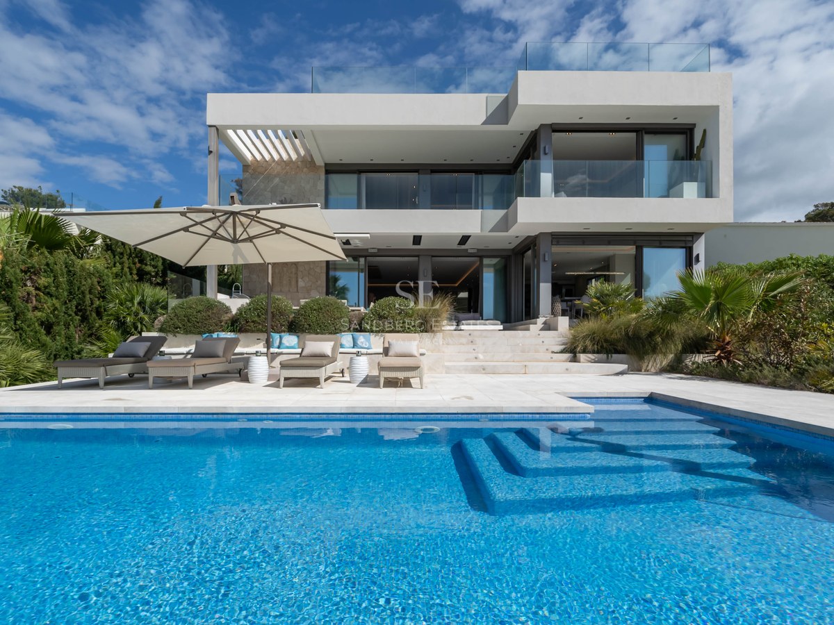 Modern two-story white villa with large swimming pool, sun loungers, and glass balconies under a blue sky.