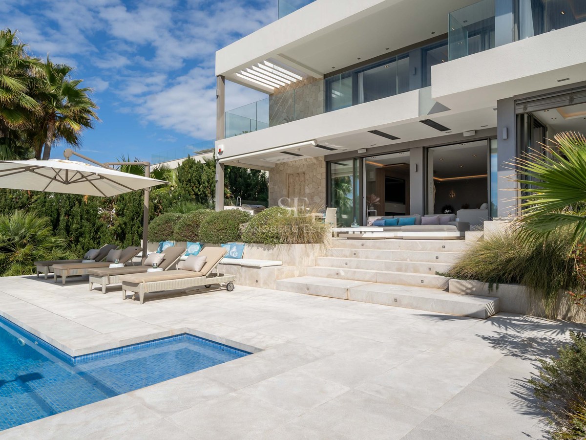 Modern villa featuring a blue tiled swimming pool, designer sun loungers, and natural stone facade under a clear sky.