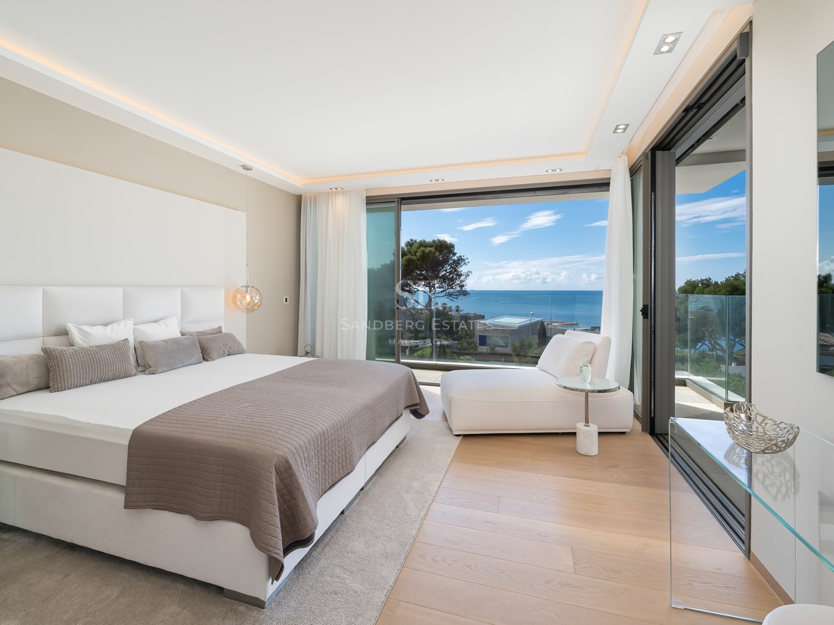 A contemporary master bedroom with a large bed, wooden floors, and floor-to-ceiling glass doors opening to a sea view.