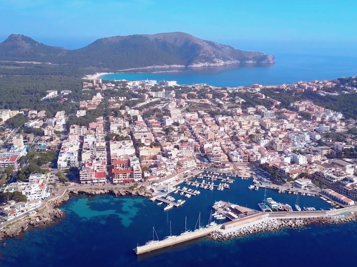Luftaufnahme eines mediterranen Yachthafens mit Booten, türkisblauem Wasser, einer Küstenstadt und Bergen im Hintergrund.