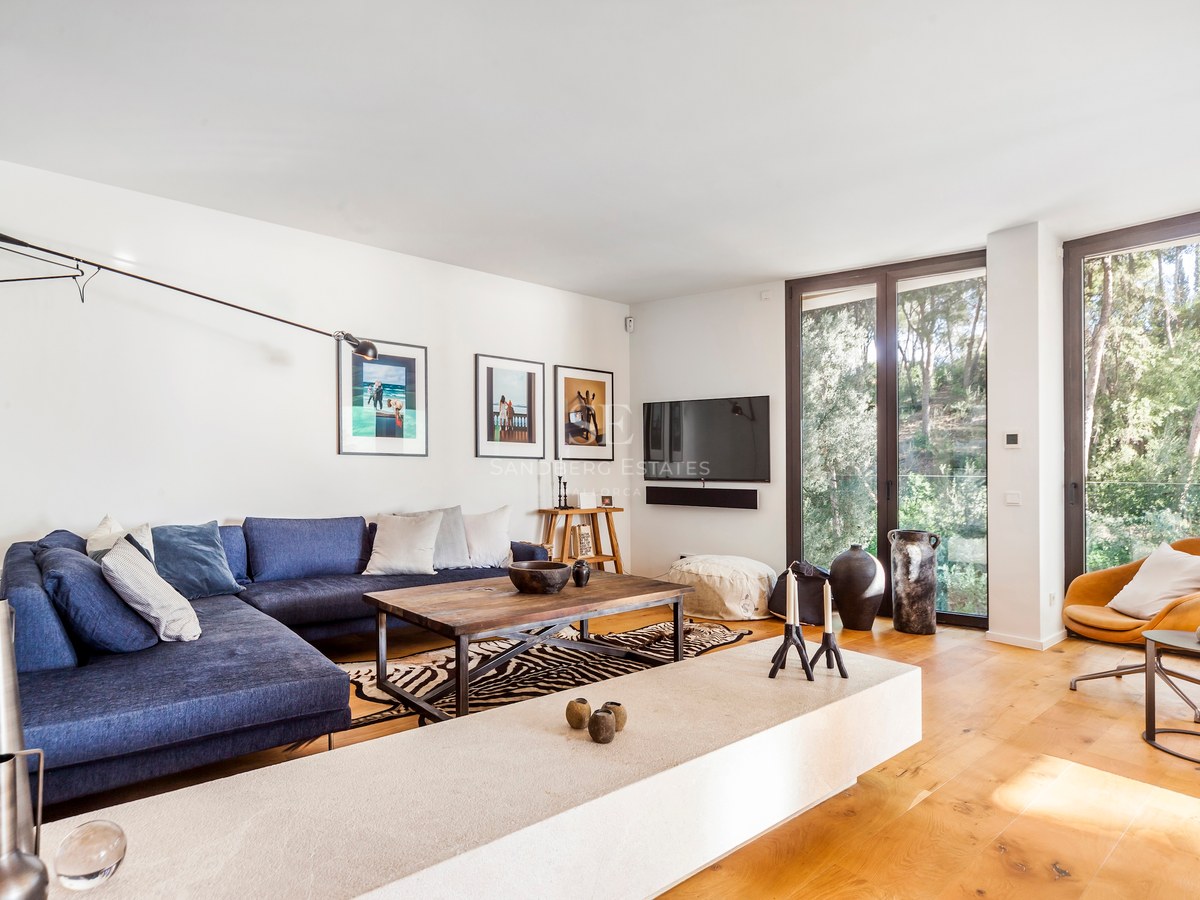 Salon moderne avec canapé bleu, parquet en chêne et grandes baies vitrées donnant sur une forêt.