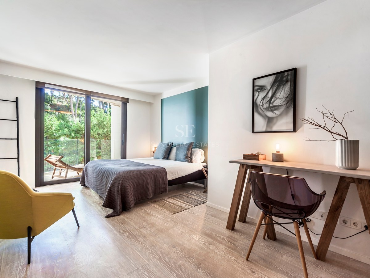 Chambre moderne avec mur sarcelle, bureau en bois, fauteuil jaune et baies vitrées ouvrant sur une terrasse verdoyante.