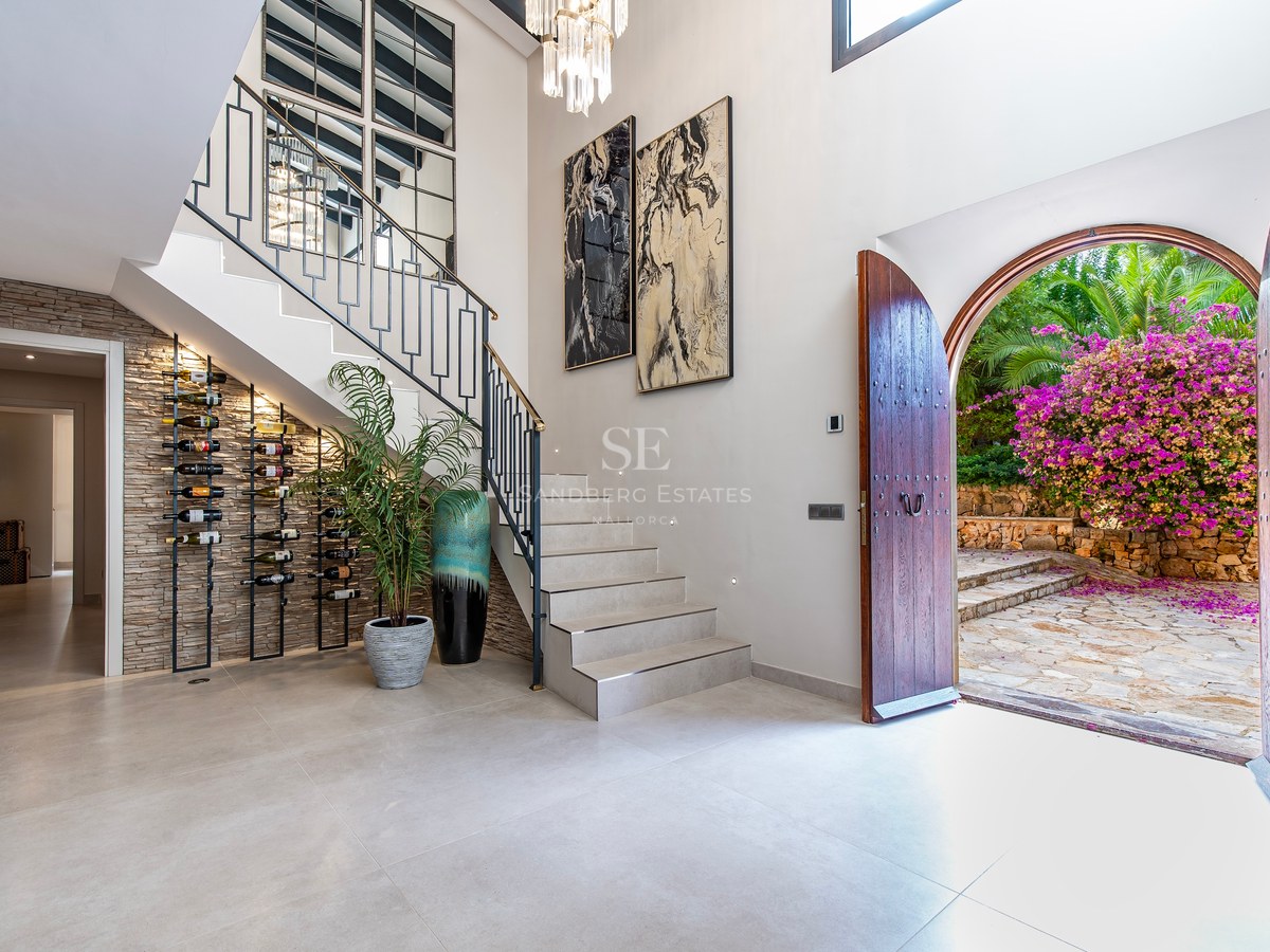 Vestíbulo de lujo con escalera moderna, botellero integrado y puertas de arco abiertas hacia un patio con buganvillas.